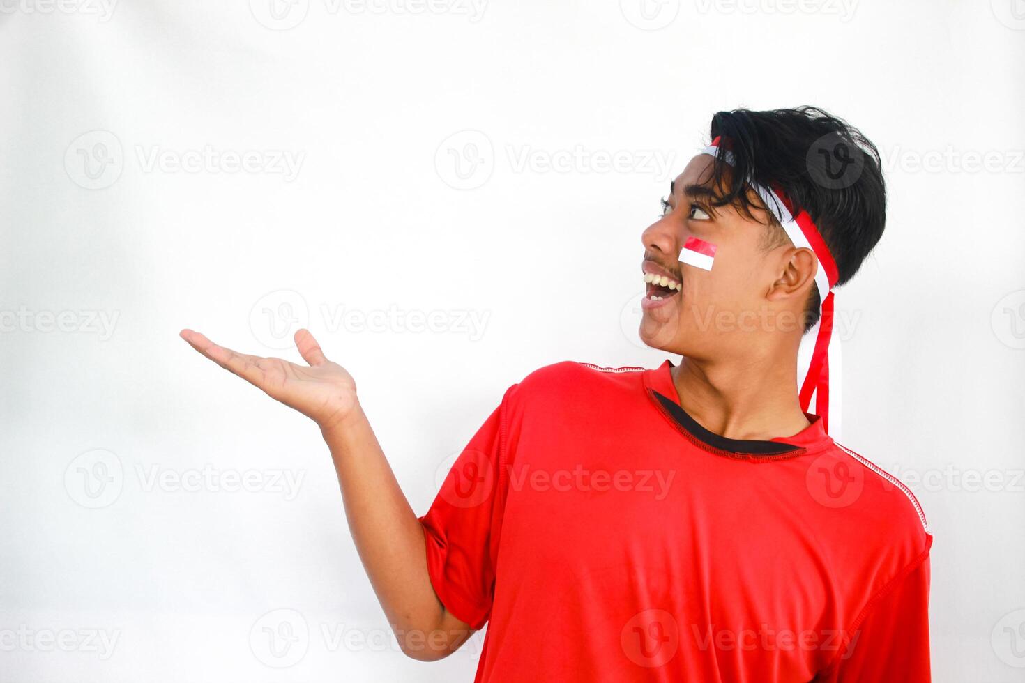 handsome young man in a red shirt and headband pointing to the copy space beside him, isolated white background, celebrating indonesia's independence day. Indonesia Independence Day concept. photo