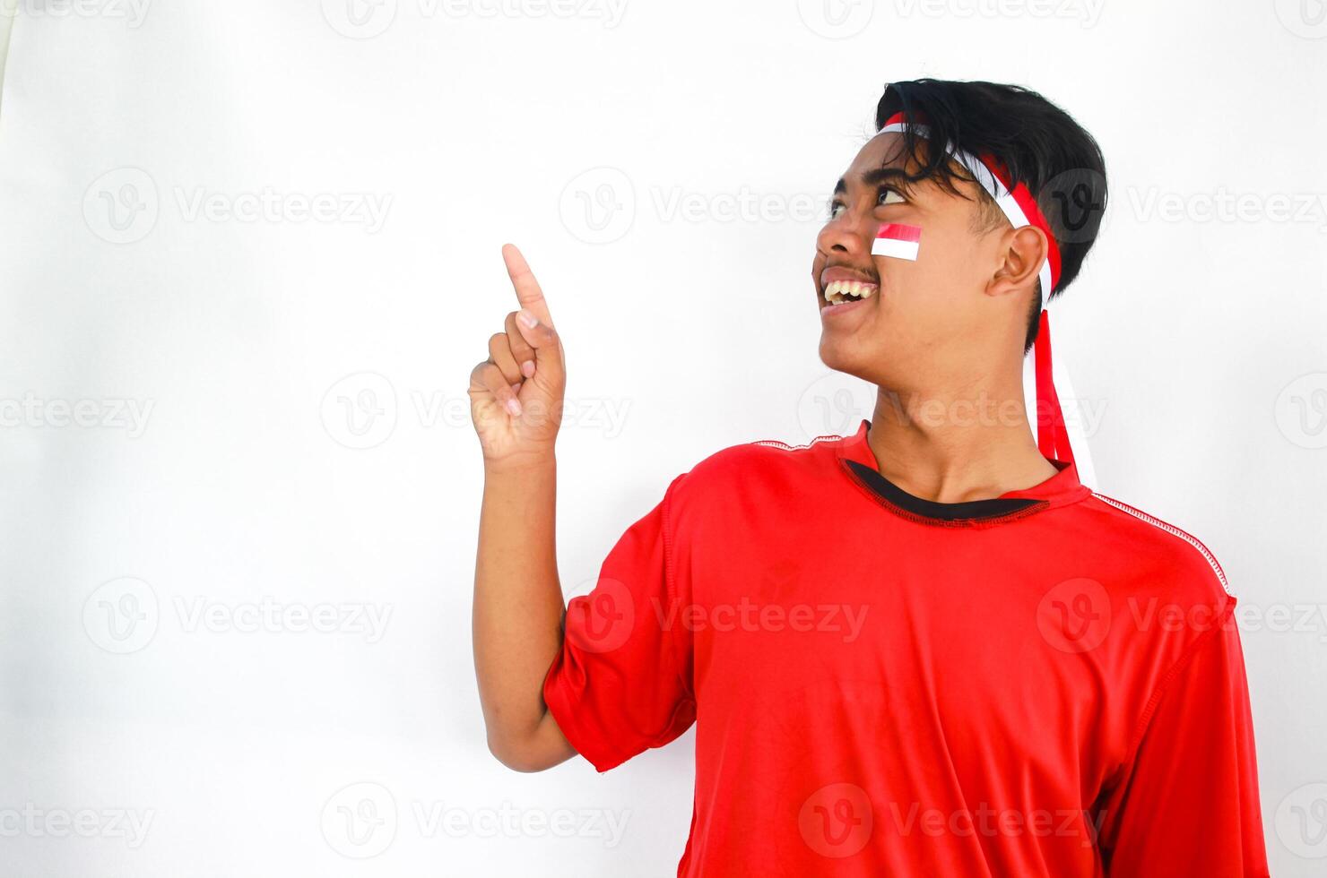 handsome young man in a red shirt and headband pointing to the copy space beside him, isolated white background, celebrating indonesia's independence day. Indonesia Independence Day concept. photo