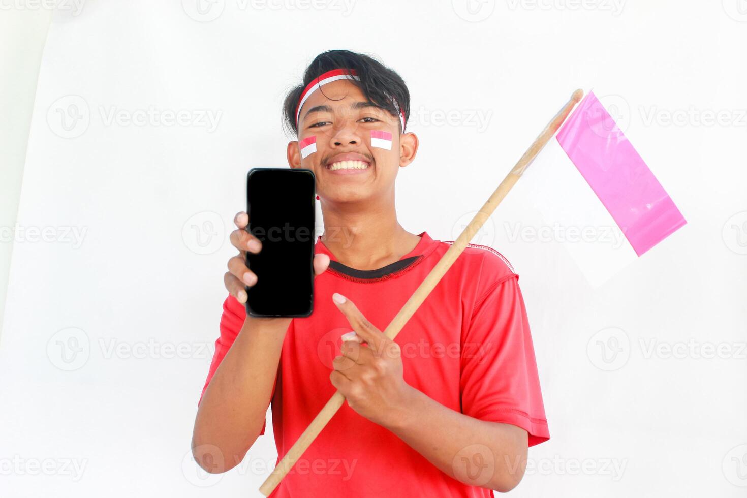 A portrait of a smiling Asian man wearing headband and showing his phone, celebrating Indonesia's independence day isolated by white background. Indonesia's independence day concept photo