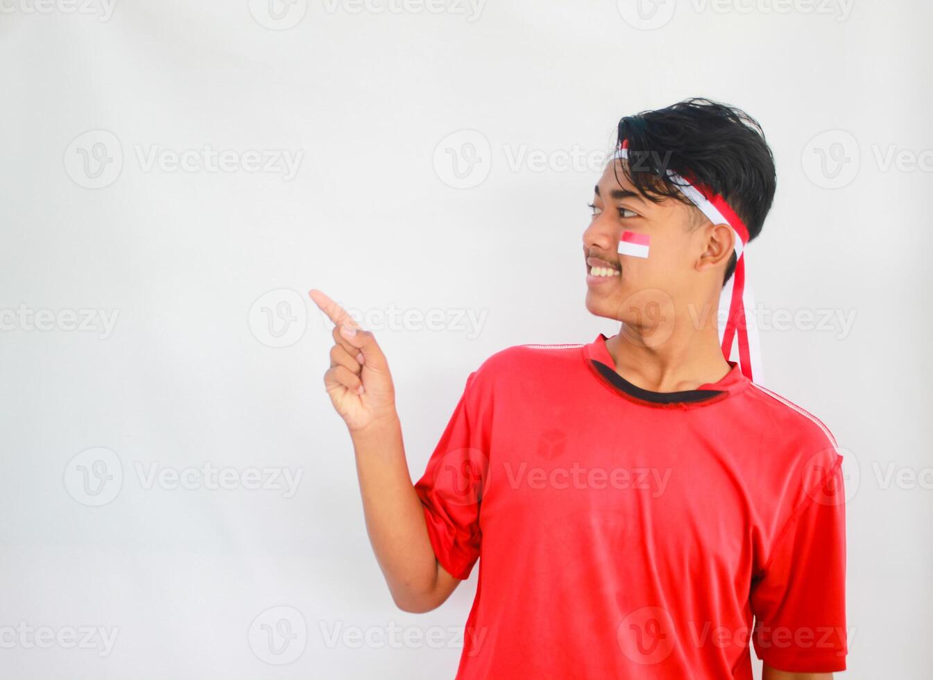 handsome young man in a red shirt and headband pointing to the copy space beside him, isolated white background, celebrating indonesia's independence day. Indonesia Independence Day concept. photo