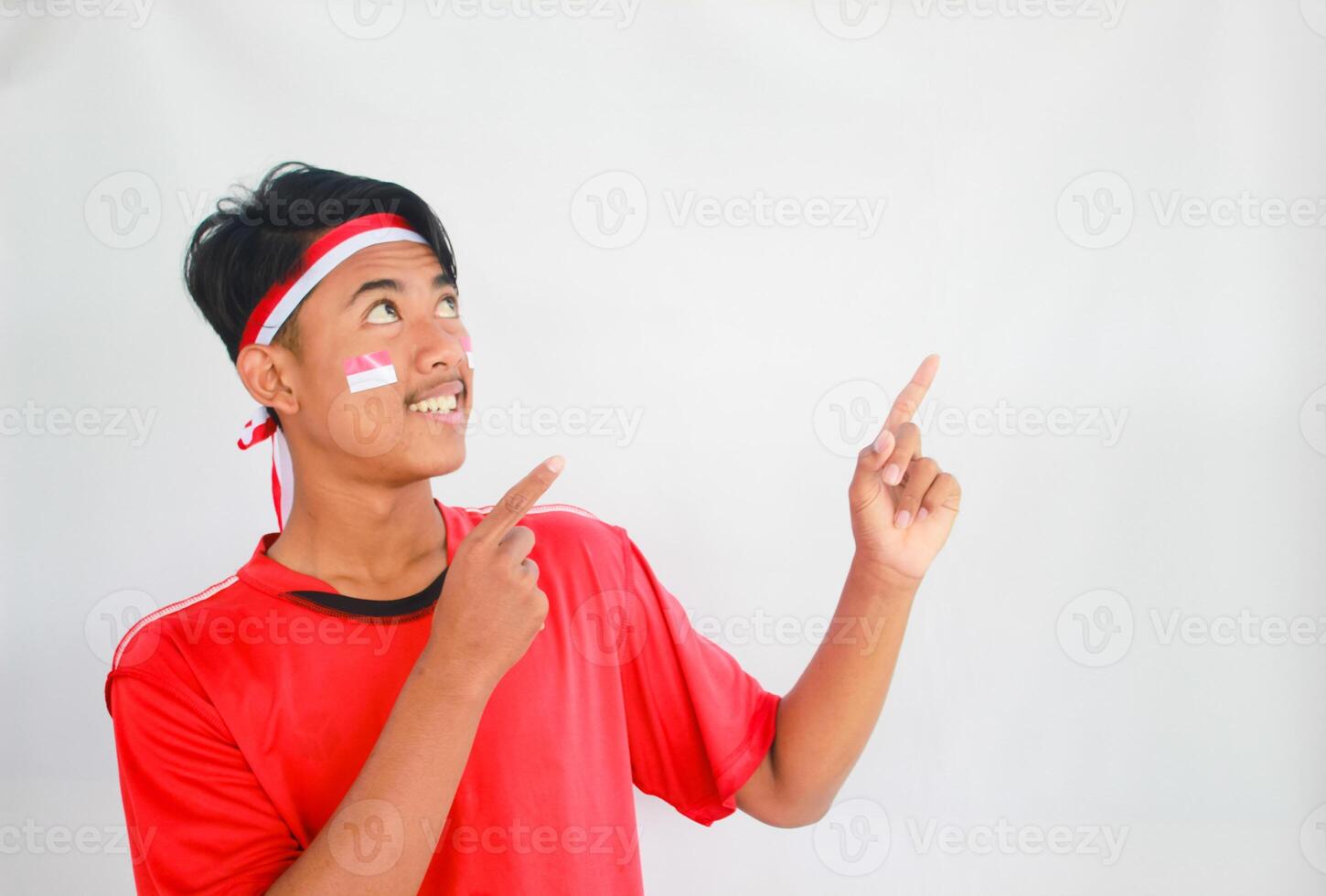 handsome young man in a red shirt and headband pointing to the copy space beside him, isolated white background, celebrating indonesia's independence day. Indonesia Independence Day concept. photo