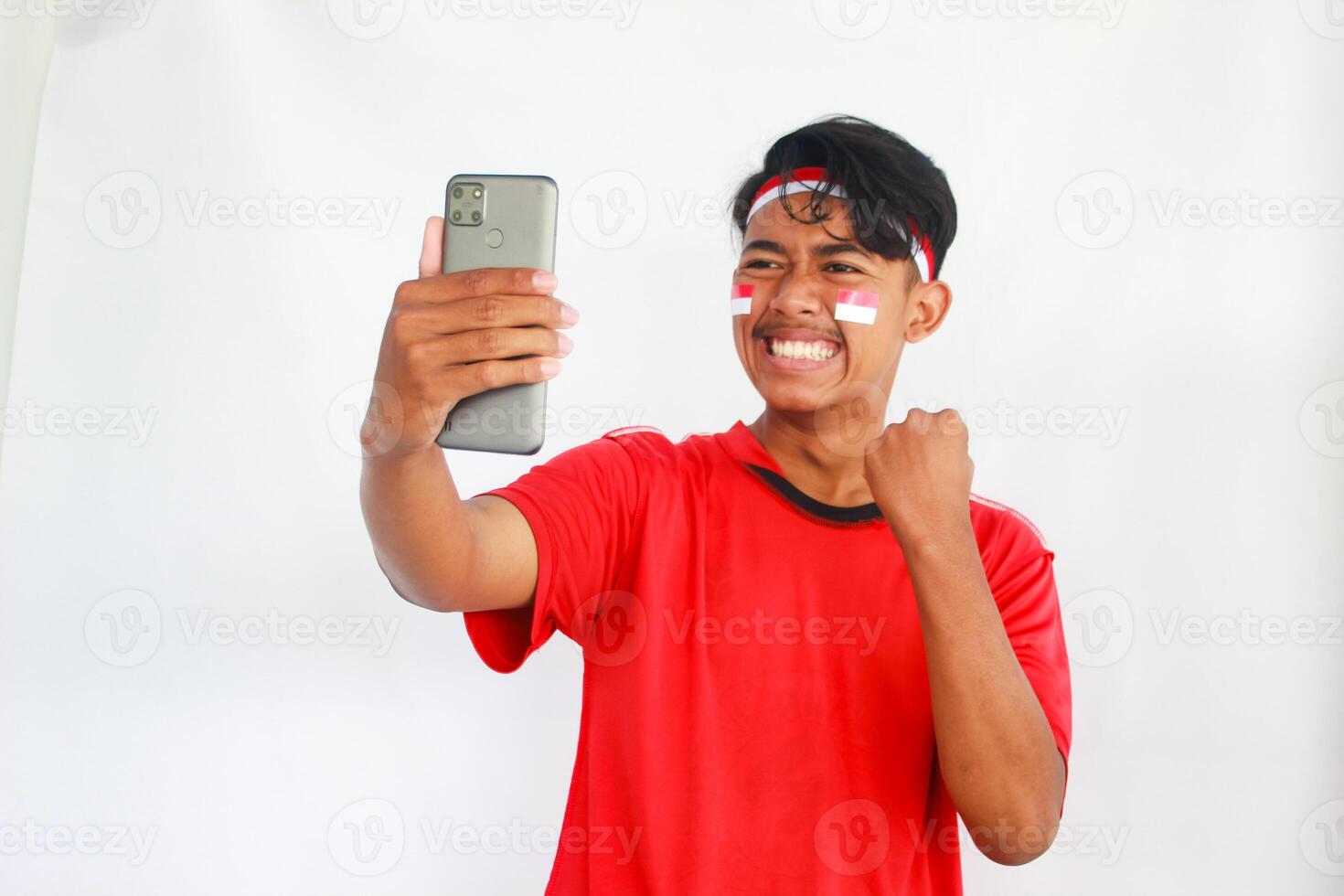 A young Asian man with a happy successful expression while holding his phone and wearing red top, flag headband and cape isolated by white background. Indonesia's independence day concept. photo