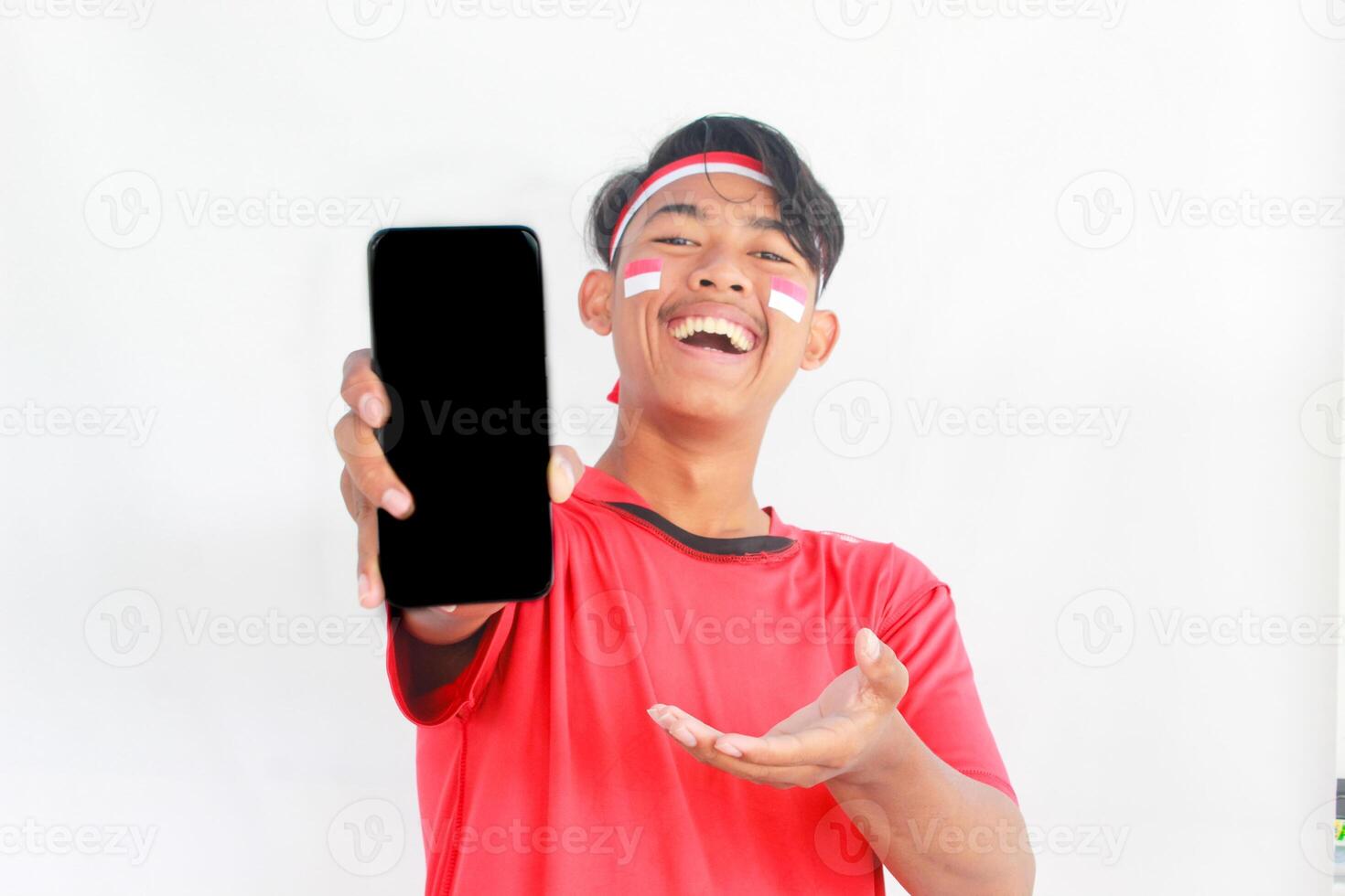 A portrait of a smiling Asian man wearing headband and showing his phone, celebrating Indonesia's independence day isolated by white background. Indonesia's independence day concept photo
