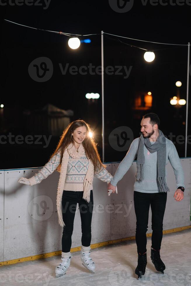 Couple on the city rink in a winter evening. Guy helping nice girl to skate on the ice in the dark night and twinkles lighting above them photo