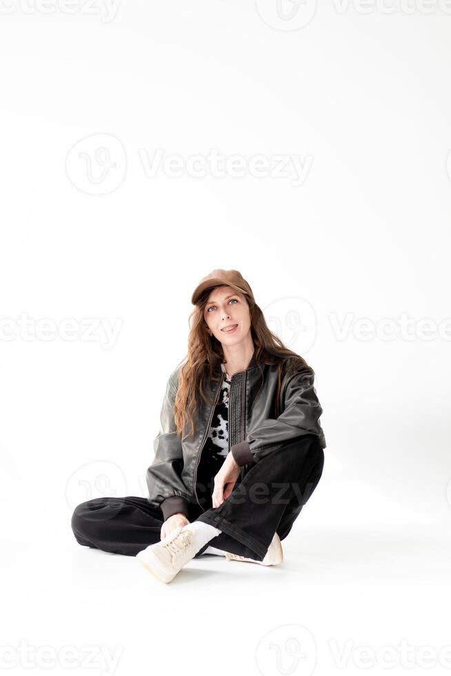 Young woman in a bomber jacket, jeans and a cap with braces poses while sitting in the studio, on a white background photo