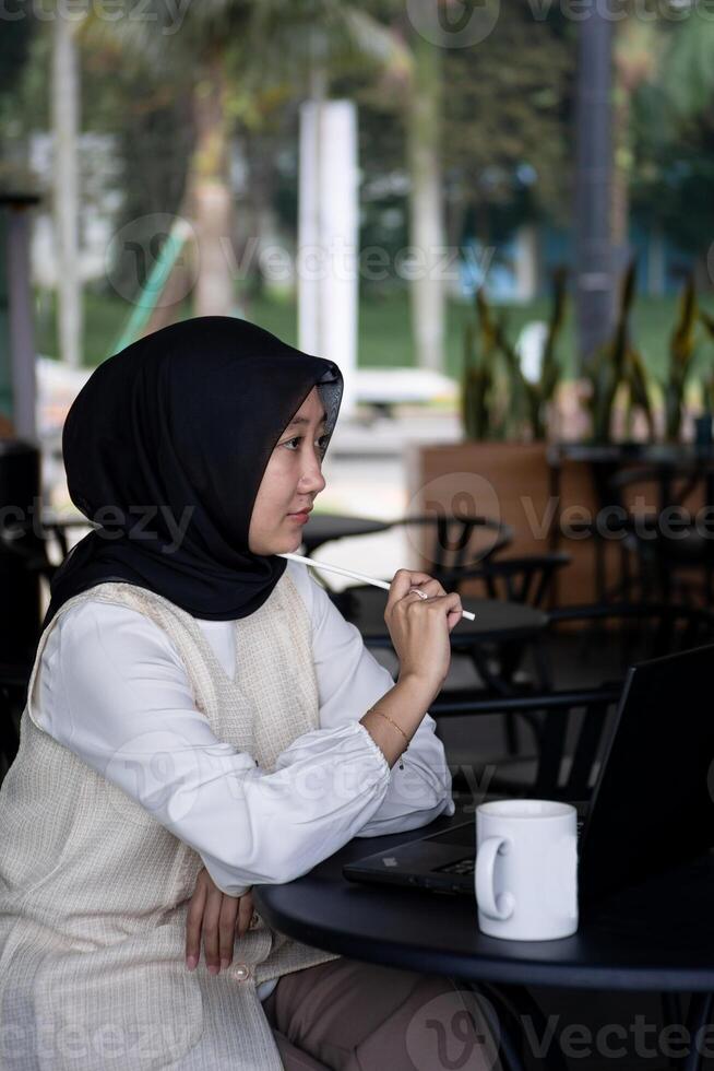 Beautiful young woman is working on a laptop and smiling, and thinking while sitting outdoors in a cafe enjoying a cup of coffee. photo