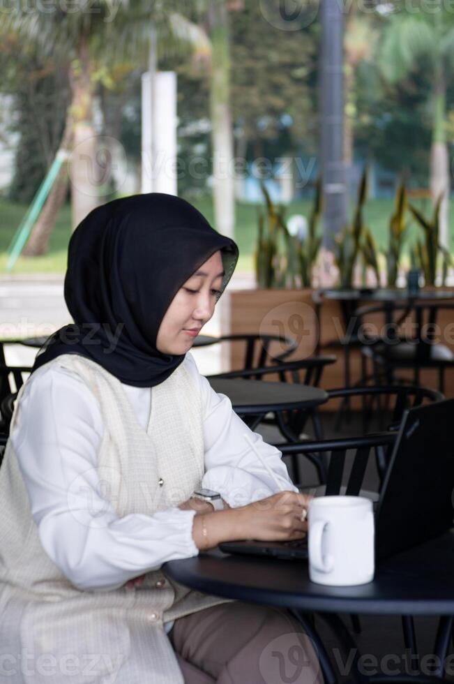Beautiful young woman is working on a laptop and smiling, and thinking while sitting outdoors in a cafe enjoying a cup of coffee. photo