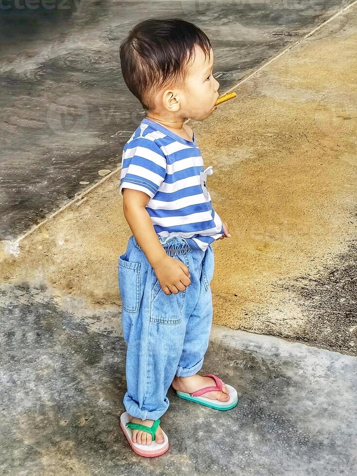 A boy in a blue striped shirt and jeans standing with a candy bar in his mouth. photo