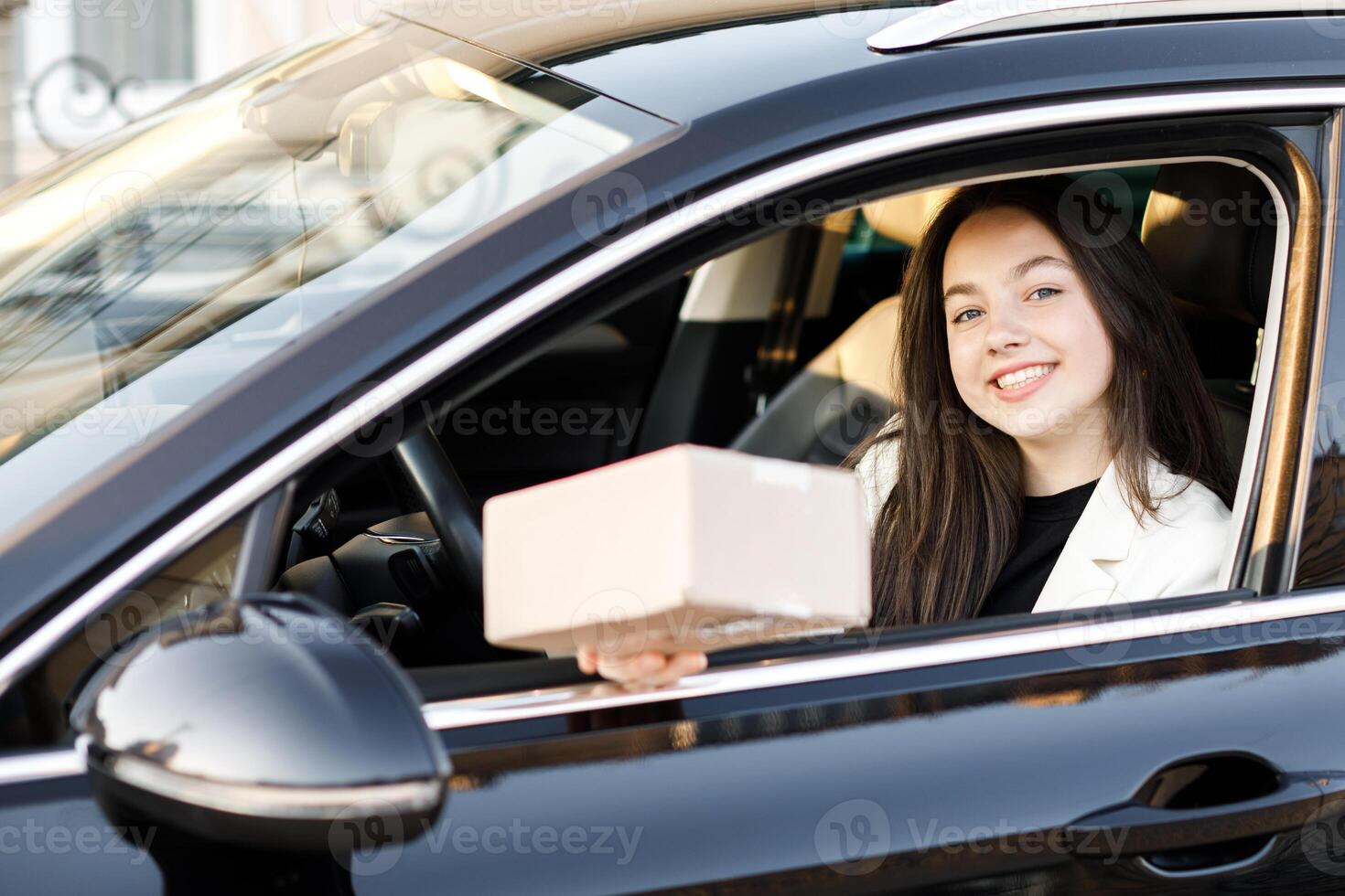 Young and happy business woman giving a parcel through the vehicle window on the go while driving a car. Concept of fast delivery on the go photo