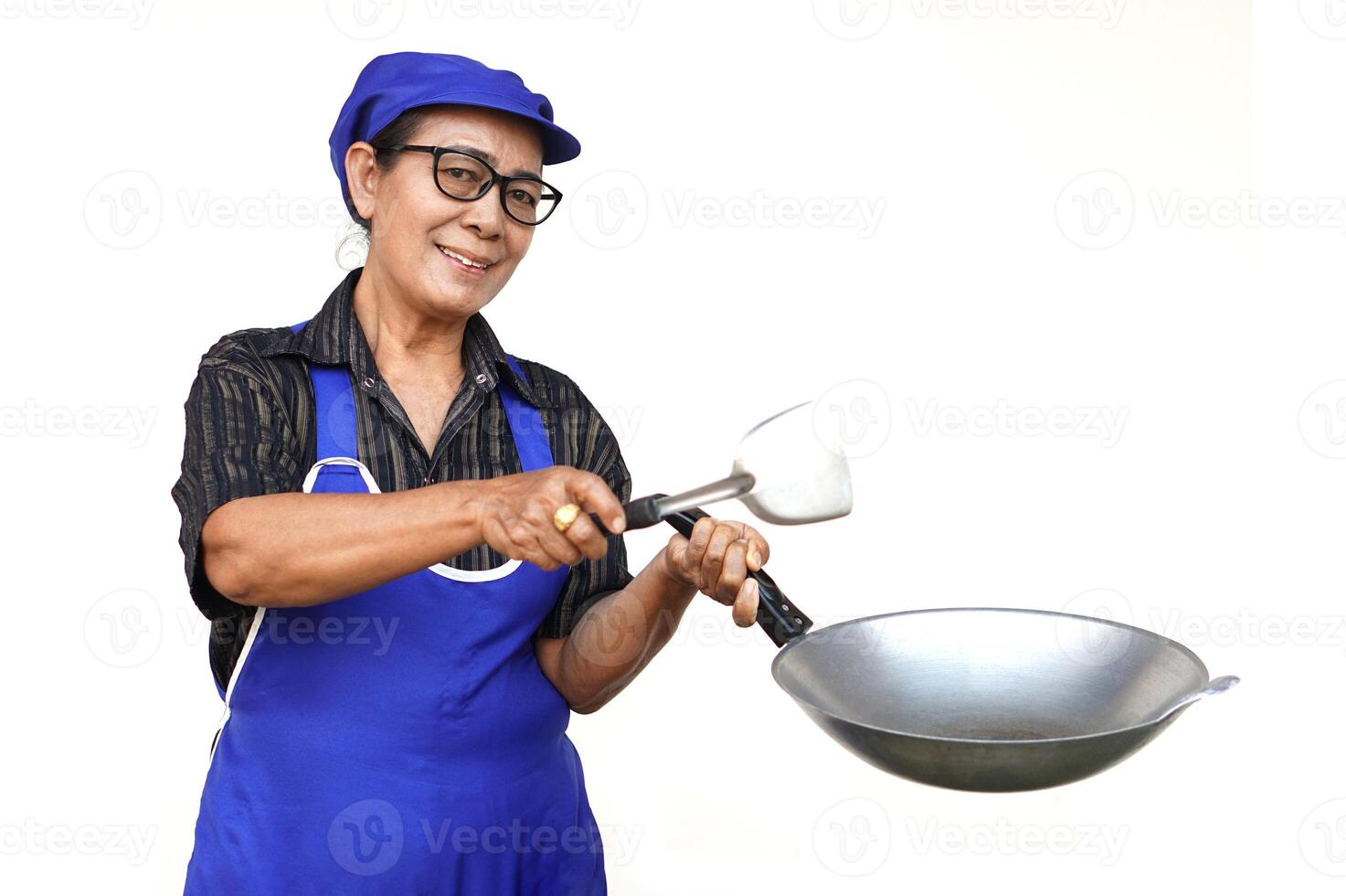 Asian senior woman, wears blue apron and cap, holds frying pan and spatula or ladle. Look at camera, smiles, feels confident isolated on white background. Concept, love cooking. Kitchen lifestyle. photo