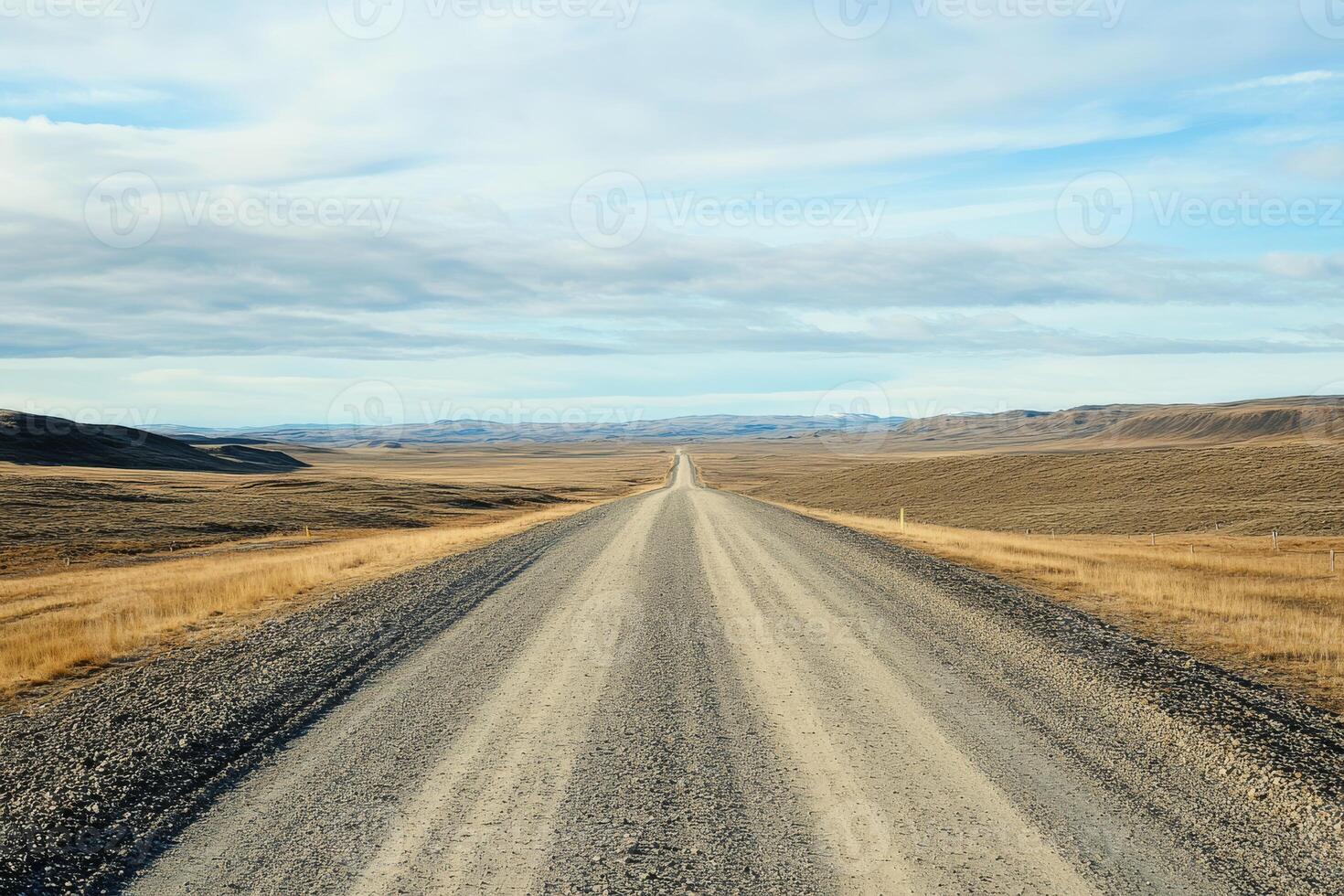 A lonely gravel road stretches out towards the distant horizon, winding its way through a barren, expansive landscape that lies beneath a dramatic, cloudy sky filled with muted light and shadows photo