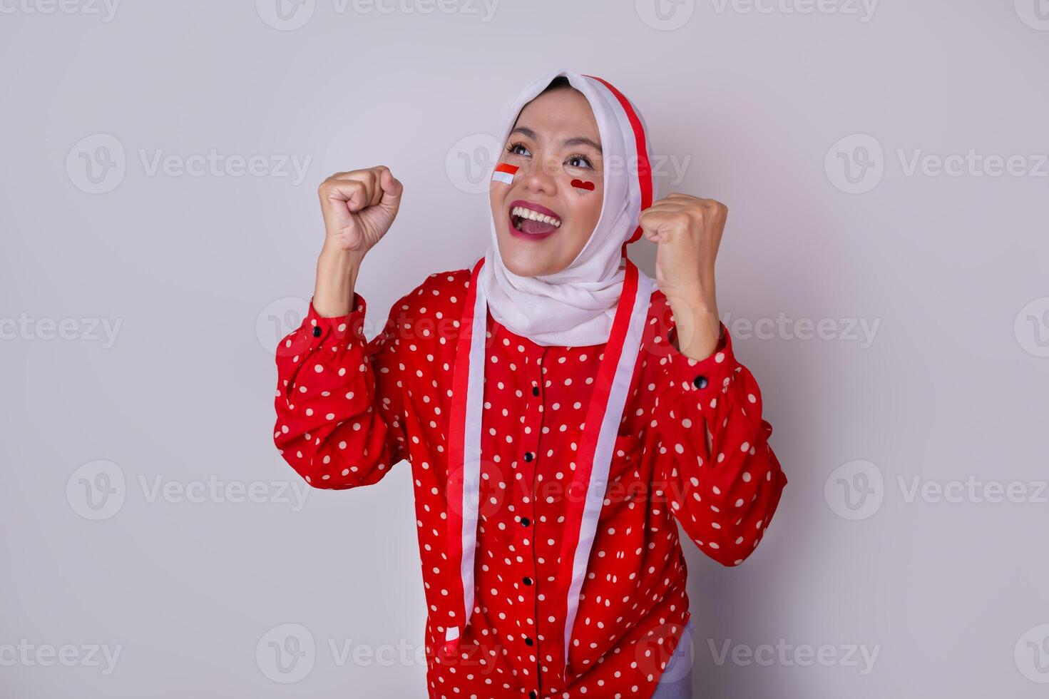 A hijab-wearing woman in a red outfit polkadot and an Indonesian flag headband poses in a celebration of Independence Day. The photo reflects patriotism and national pride.