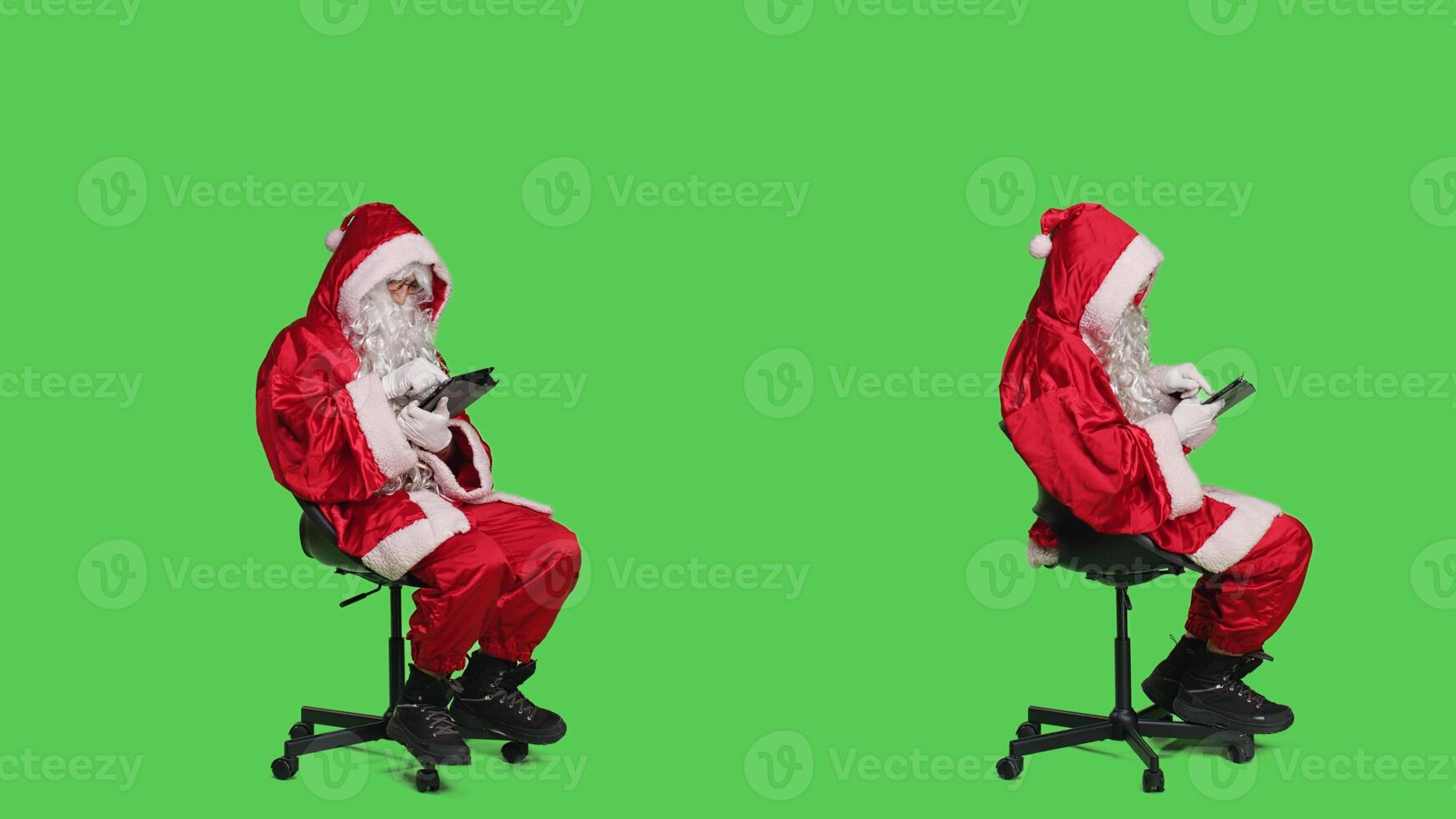 Modern santa works on digital tablet during christmas holiday celebration, looking on online webpage while he sits on chair. Young man against full body greenscreen backdrop using device. photo