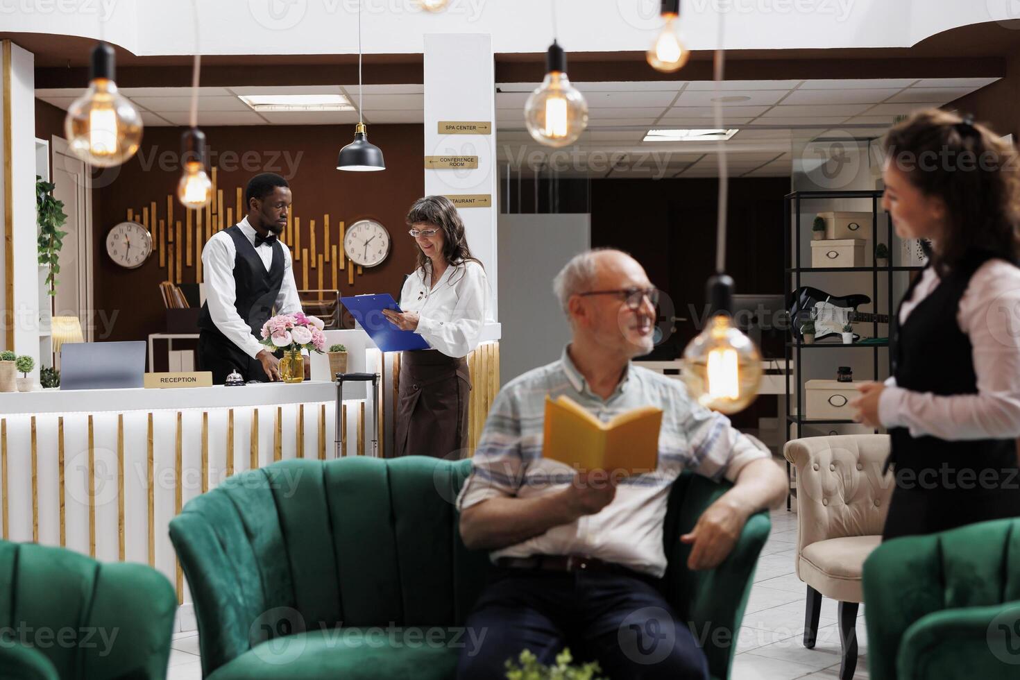 Senior female guest signs check-in papers at hotel reception front desk. Friendly male concierge assists old woman as she fills out reservation forms while caucasian waitress helps old man in lounge. photo