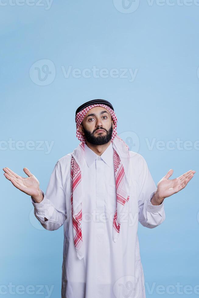 Confused muslim man posing in studio with open arms, expressing hesitation gesture. Arab person wearing traditional clothes spreading hands and shrugging shoulders portrait photo