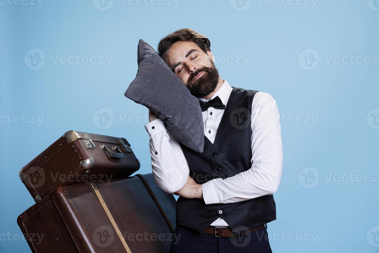 Tired bellboy posing with pillow on camera, expressing burnout and feeling sleepy at professional classy job. Hotel concierge taking quick nap after being overworked, exhausted doorman. photo