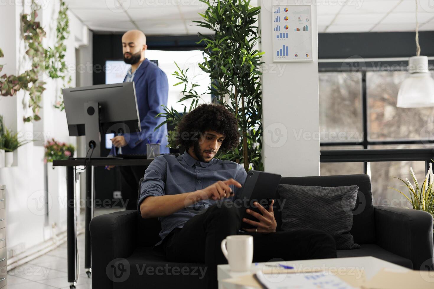 Start up entrepreneur checking project presentation on digital tablet in office. Young arab businessman sitting on couch and using portable gadget to send message in social media photo