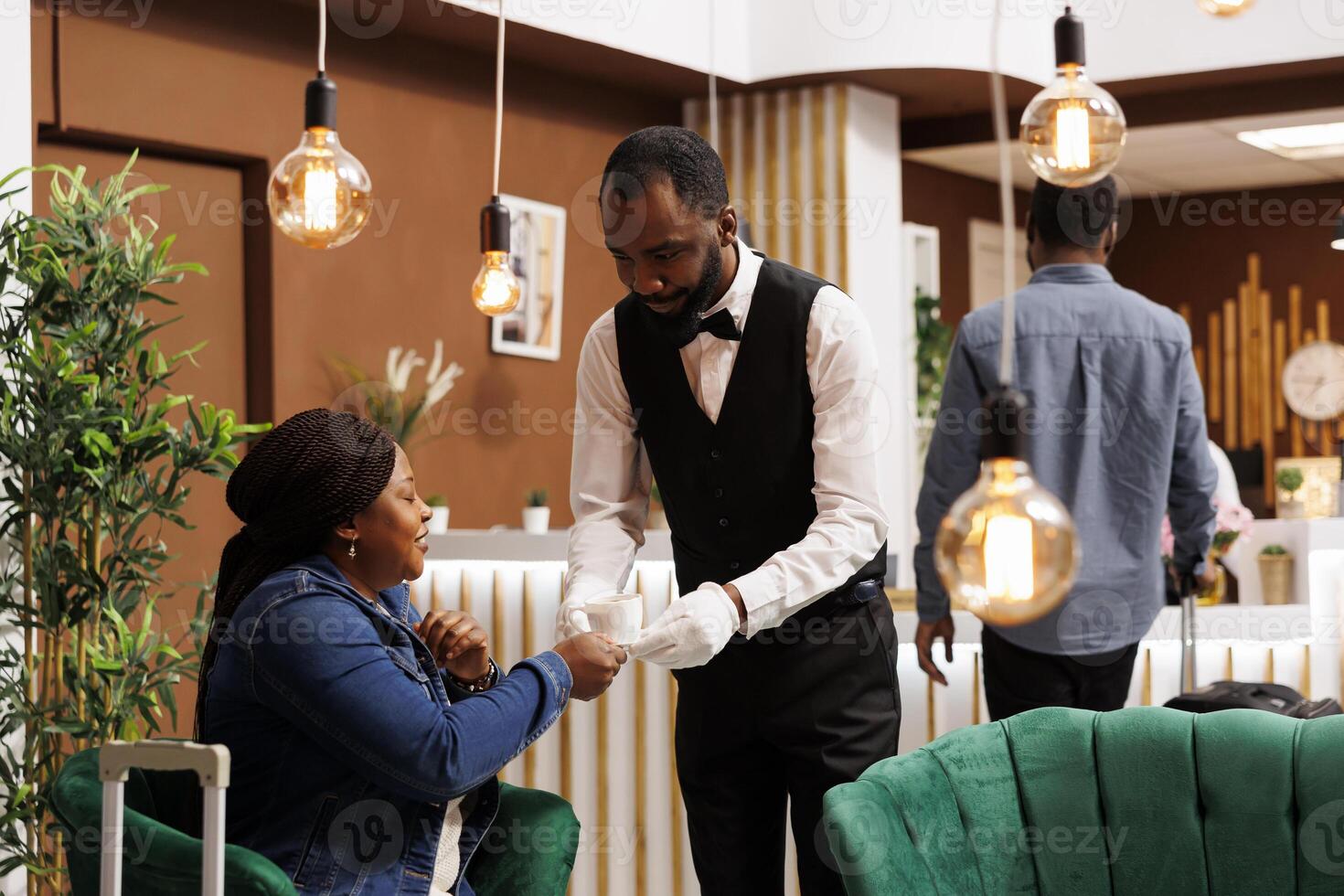 Smiling friendly African American guy waiter holding cup of fresh brewed coffee, serving it to happy female hotel guest waiting for check-in at lobby. Caffeine, travel and jet lag concept photo