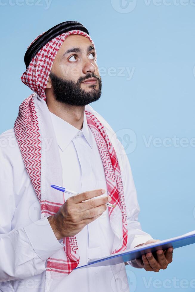 Thoughtful man wearing muslim traditional clothes looking upwards while writing in clipboard. Arab person dressed in islamic headscarf and thobe holding pen and taking notes in checklist photo