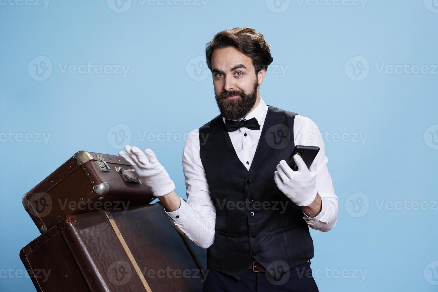 Professional bellman holds smartphone in hand and popsing next to trolley bags, portraying luxurious hospitality industry occupation. Young man in formal attire using mobile phone webpages. photo