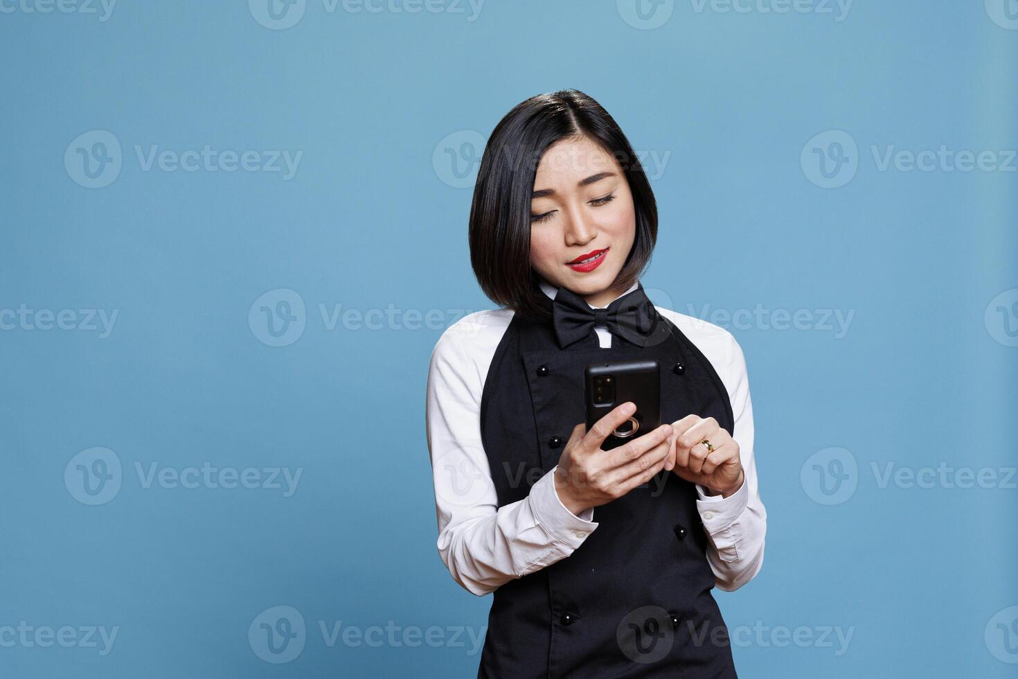 Smiling asian waitress wearing uniform texting on smartphone, managing catering service online. Restaurant woman receptionist typing message on mobile phone, posing in studio photo