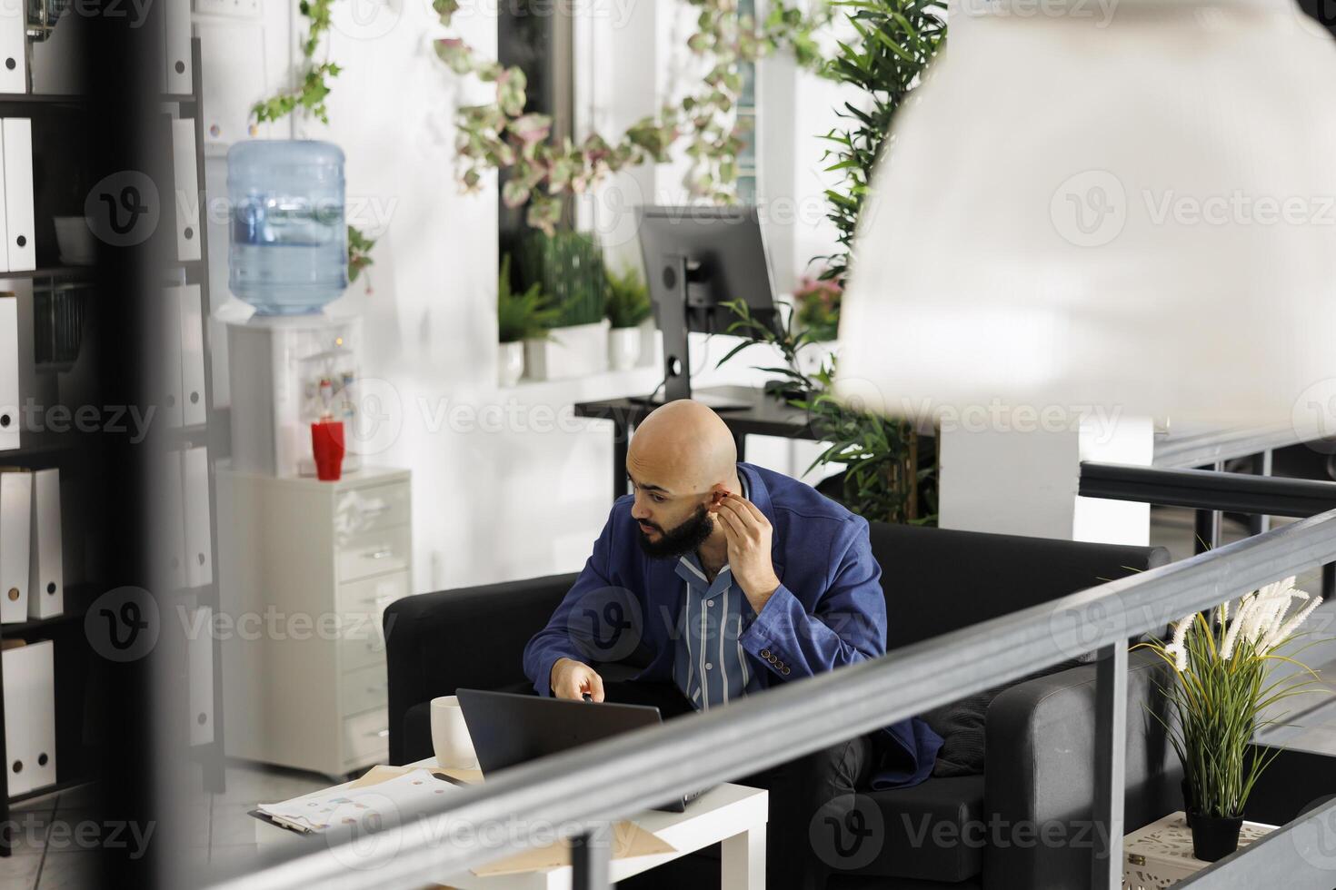 Arab corporate employee putting in earbuds to answer ceo videocall on laptop while working in startup business office room. Professional wearing earphones to talking online in green open space photo