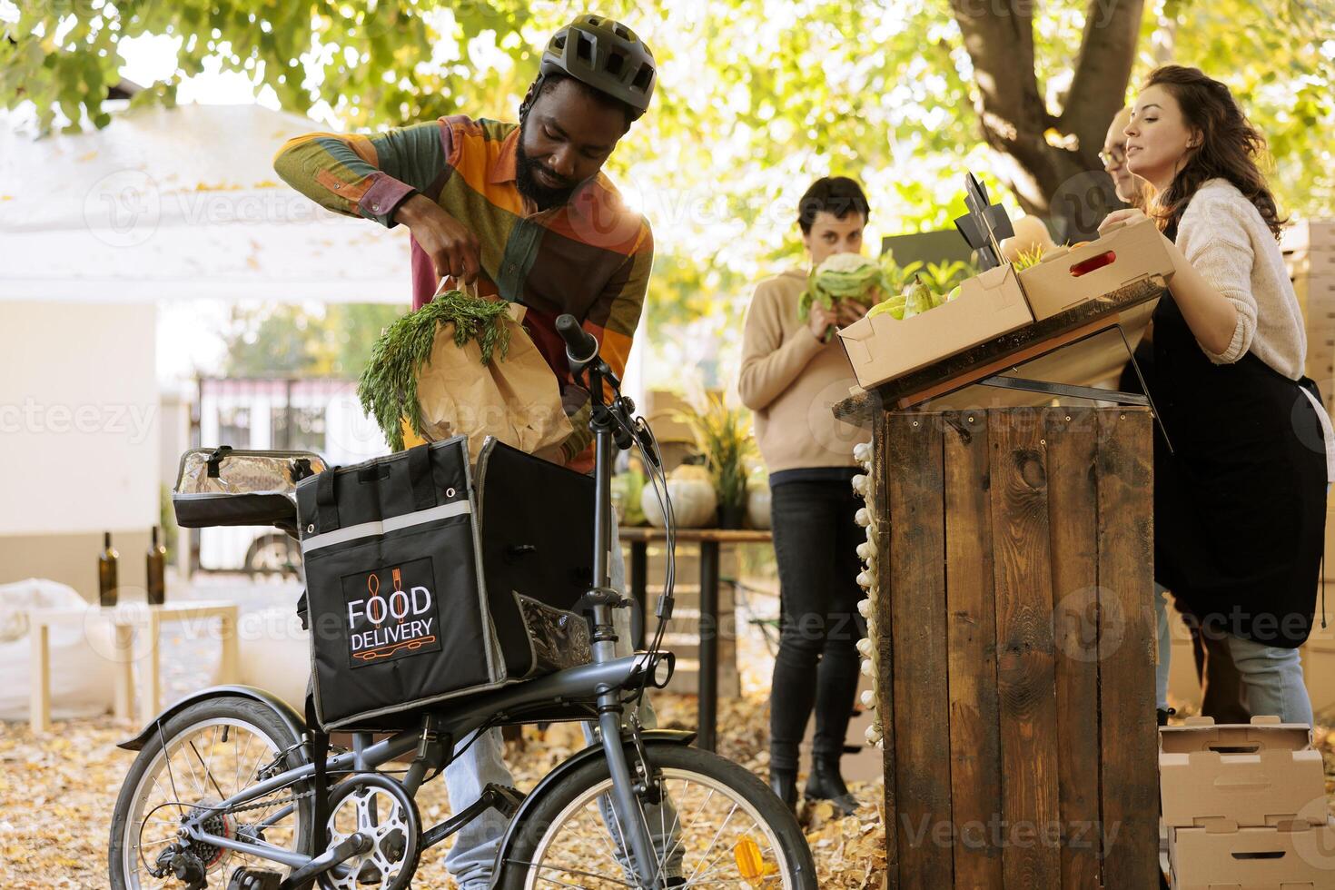 Black man is putting bio organic produce in a thermal bag, taking an order from the farmers market, and riding his bike. African courier delivering farm fresh fruits and vegetables to customers. photo