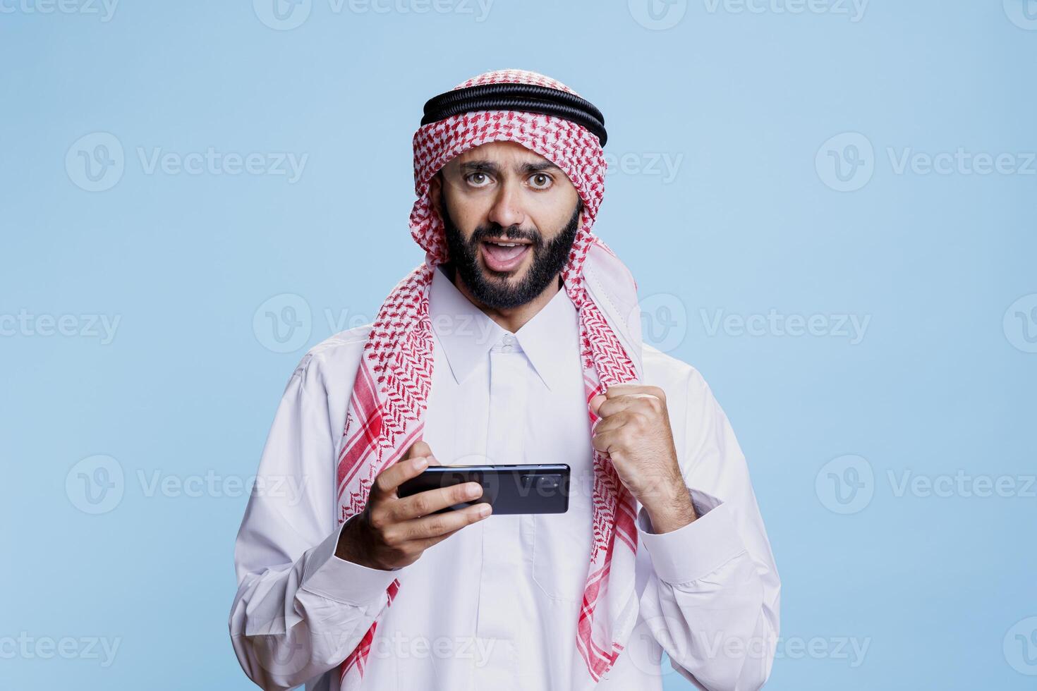 Excited arab in traditional clothes winning game on smartphone and looking at camera with clenched fist. Muslim person showing victory concept while playing videogame on mobile phone portrait photo