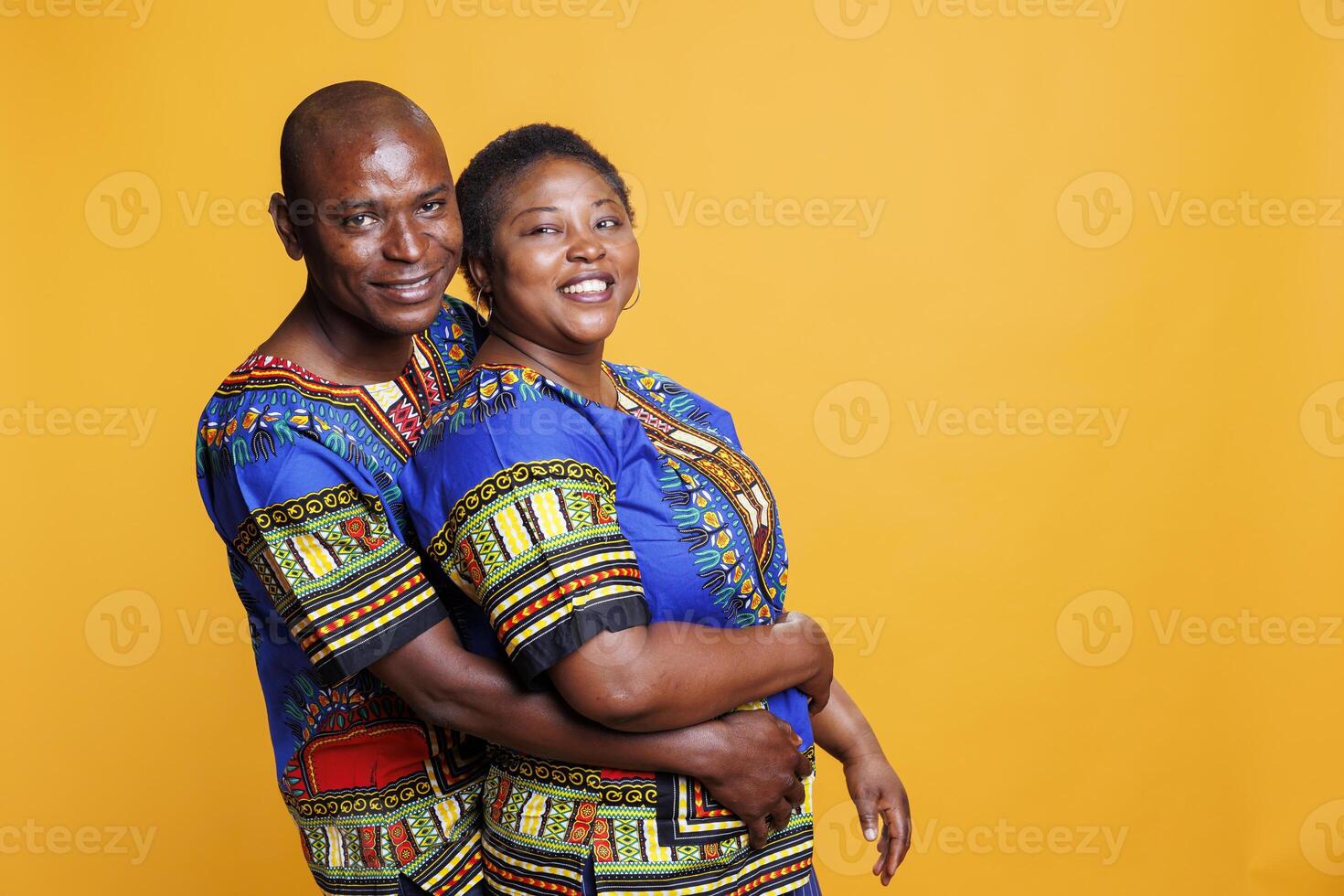 Smiling husband hugging wife from back while posing for portrait on studio orange background. Happy man and woman couple embracing and showing love while looking at camera photo