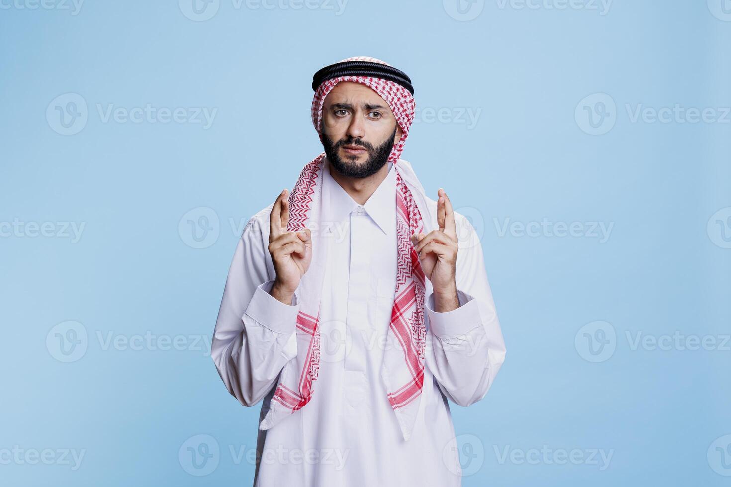 Man wearing cultural muslim clothes crossing fingers with superstitious belief and hopeful expression. Arab dressed in traditional thobe and ghutra standing with making wish and good luck gesture photo