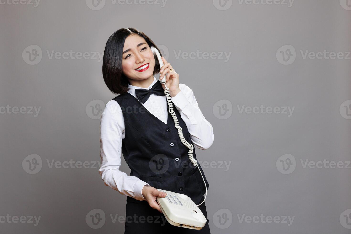 Smiling attractive asian cafe waitress speaking on landline phone, having conversation with coworker. Hospitality service cheerful friendly receptionist talking with client on telephone photo