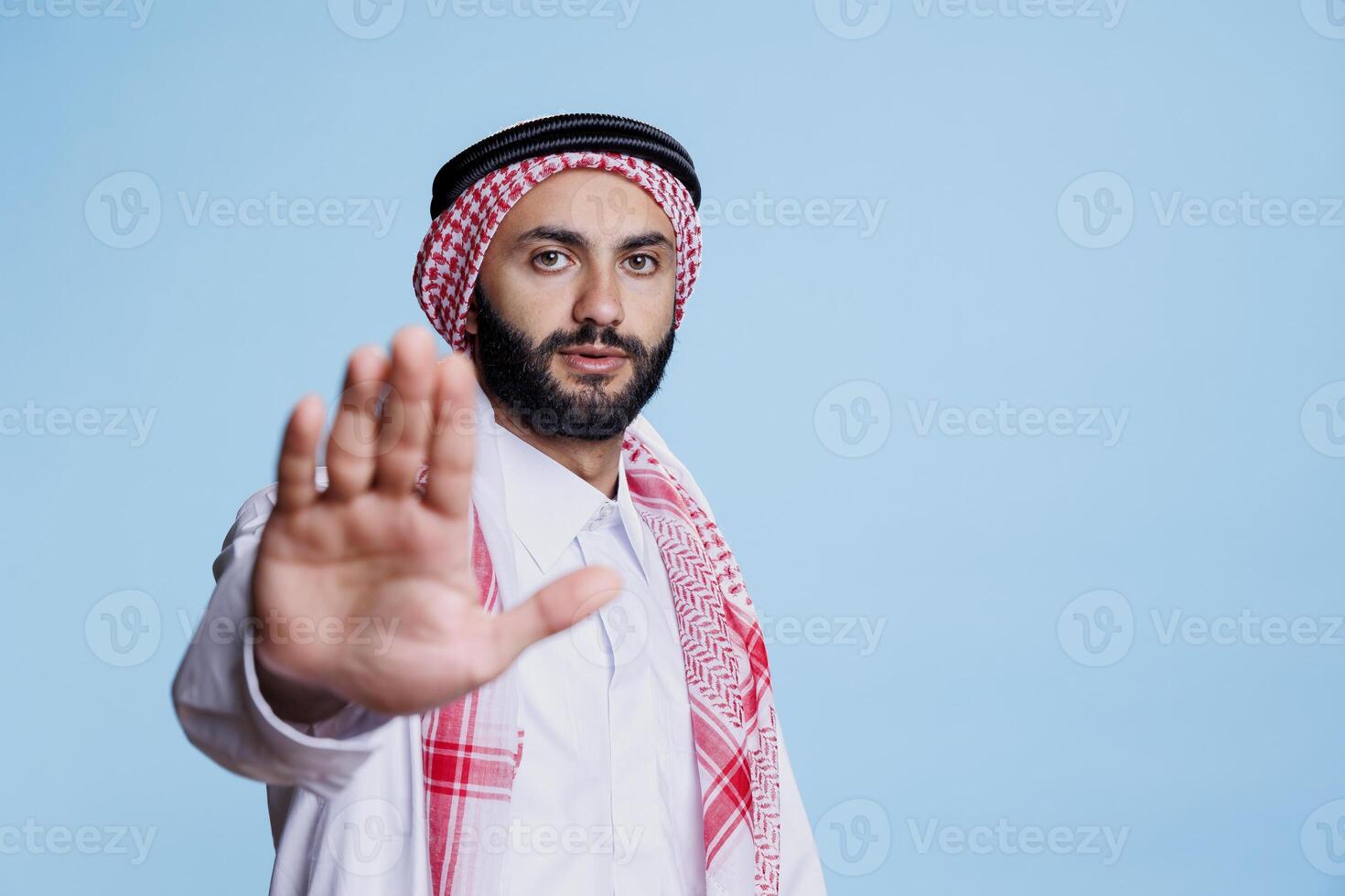 Serious man dressed in muslim thobe and headscarf making stop gesture with arm portrait. Arab person wearing traditional clothes making with prohibition with hand and looking at camera photo