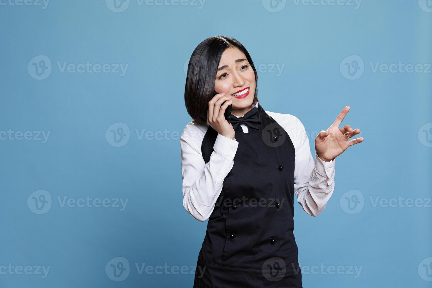 Smiling asian waitress in uniform engaging in friendly conversation on smartphone. Cheerful young attractive woman receptionist chatting on mobile phone and making gestures photo