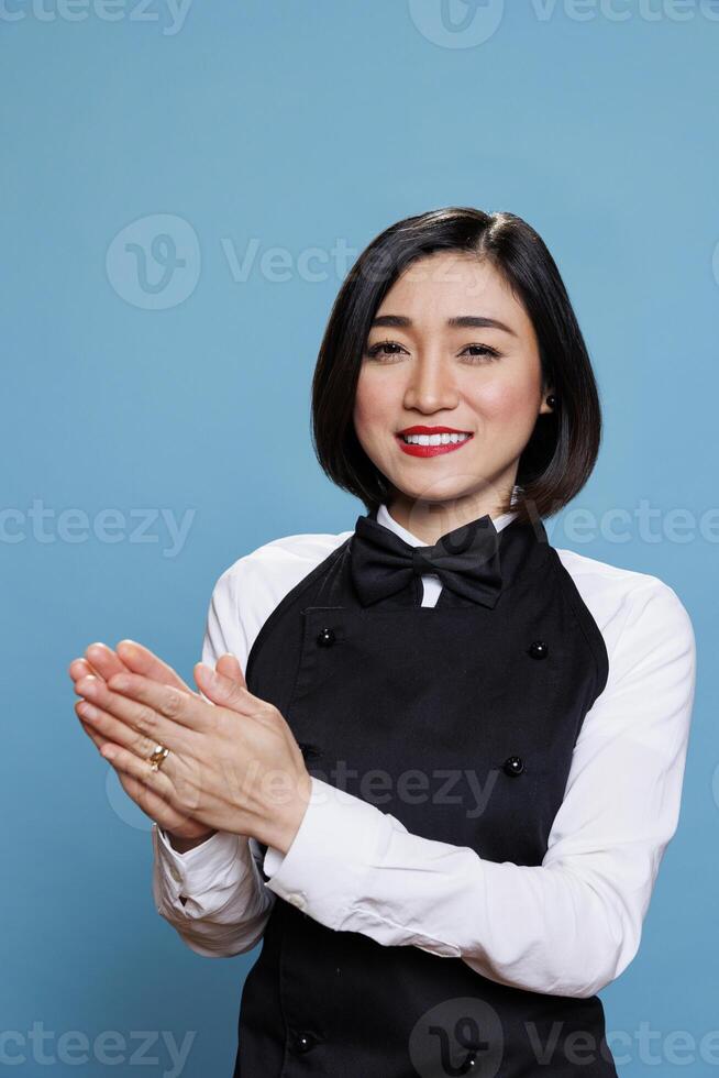 Restaurant smiling asian waitress applauding, encouraging with positive gesture and looking at camera. Cheerful cafe young woman worker in uniform making applause portrait photo