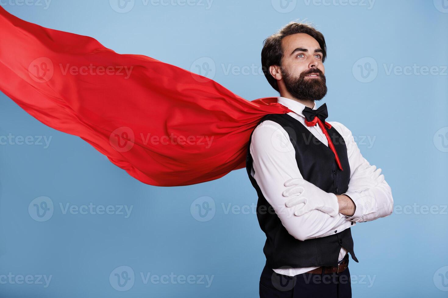 Powerful man in red hero cape posing, showing determination and force to protect people. Smiling strong adult acts as an inspiration or superhero, stands against blue background. photo