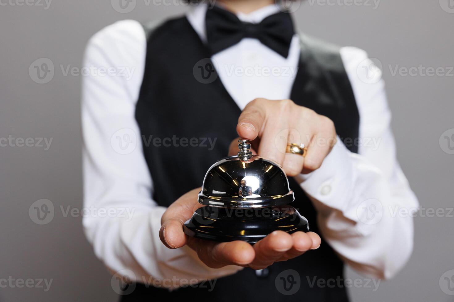 Waitress in uniform holding dining bell in hands and ringing while posing in studio closeup. Catering service staff pressing order notification alert with finger on gray background photo