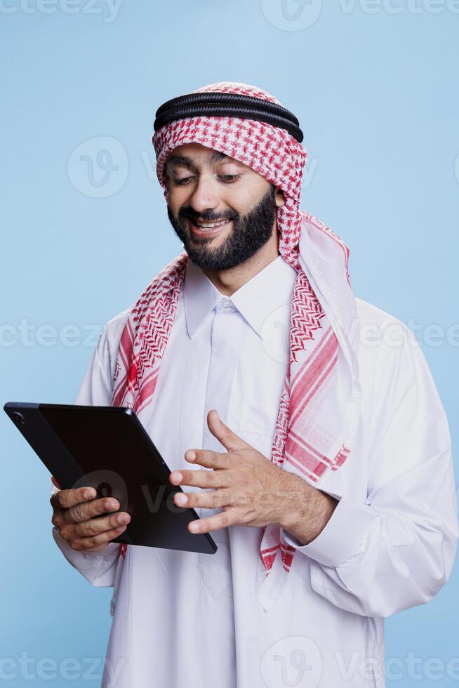 Smiling man wearing muslim thobe and headdress using digital tablet with cheerful expression. Arab person in traditional clothes scrolling through internet page on portable gadget photo