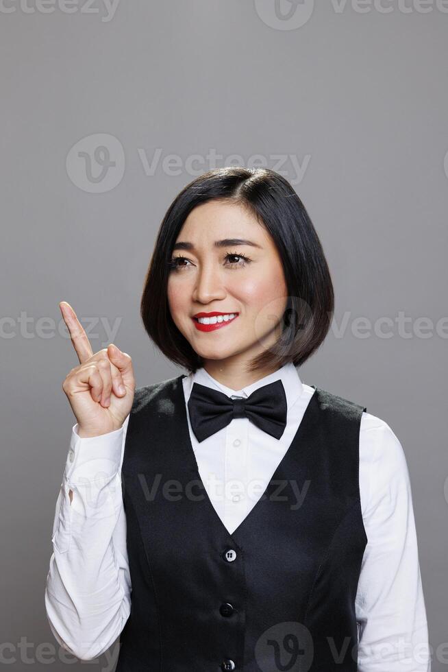 Cheerful asian woman wearing waitress uniform directing upwards with finger and posing in studio with joyful facial expression. Restaurant smiling receptionist pointing up photo