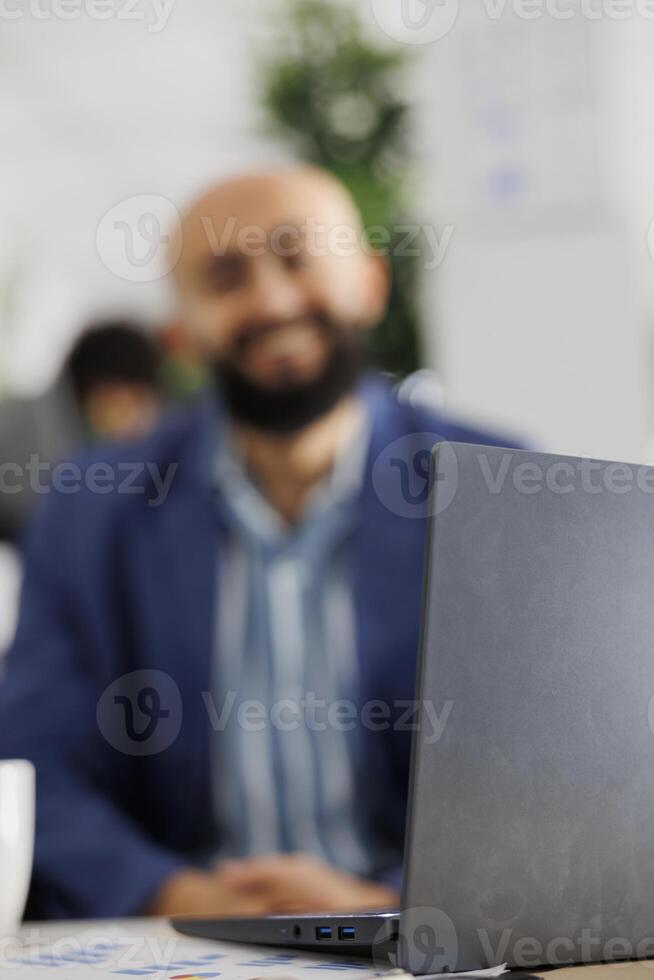 Blurred company employee working on laptop in business office. Smiling arab man start up entrepreneur using portable computer at workplace in coworking space and looking at camera photo