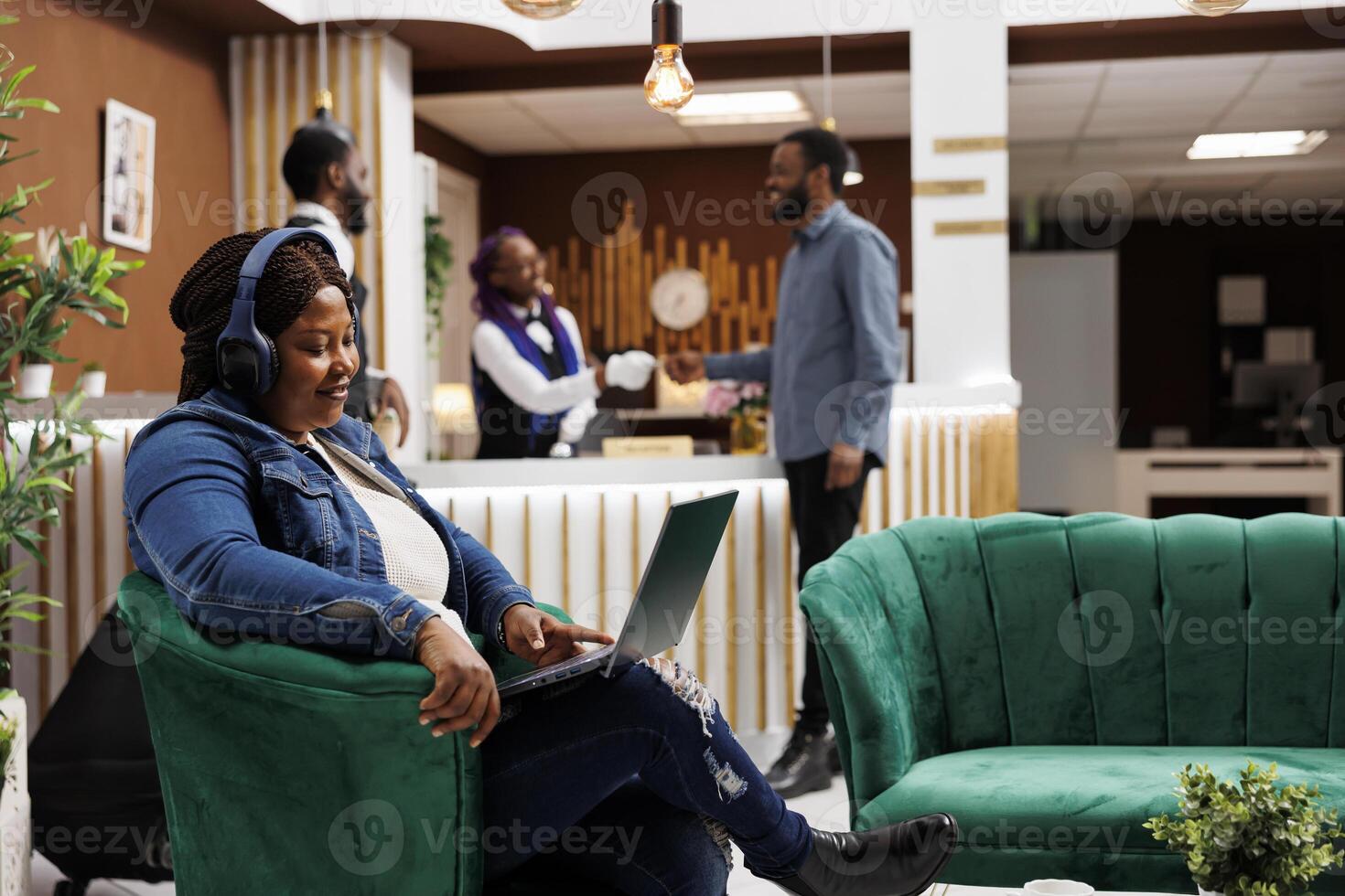Smiling happy African American woman traveler resting in hotel lobby using headphones and laptop, wathing movie online or talking with family via call. Tourist using free wifi hotspot at resort photo