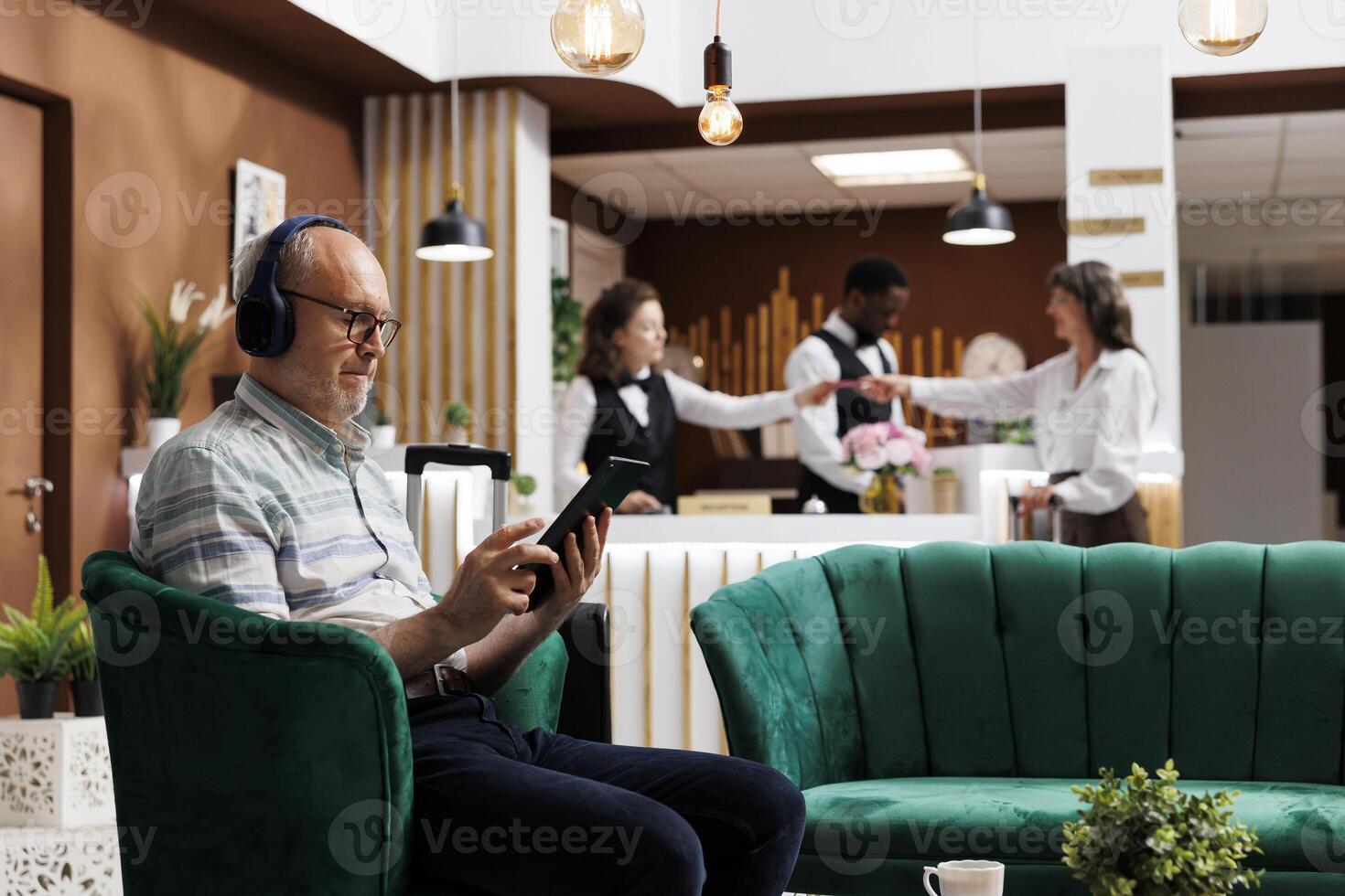 Female client checks-in at hotel reception while eldery male traveler browses and does room booking using tablet. Retired senior man with headphones, using his device for online check-in. photo