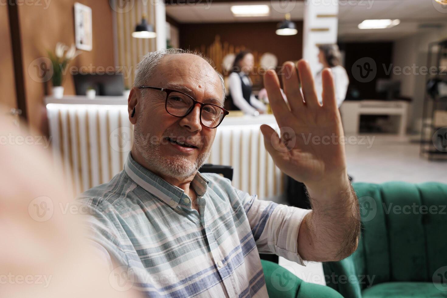 Smiling elderly man in hotel lounge area takes a selfie on his mobile phone while staff assists other guest in background. Retired senior tourist capturing fun holiday vacation on smartphone. photo