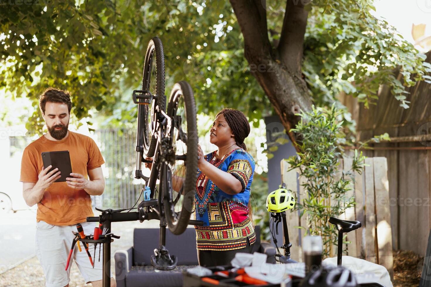 African american woman secures bike on repair-stand while caucasian man uses phone tablet to research information for repairing. Two sporty persons outside using digital device for mending bicycle. photo