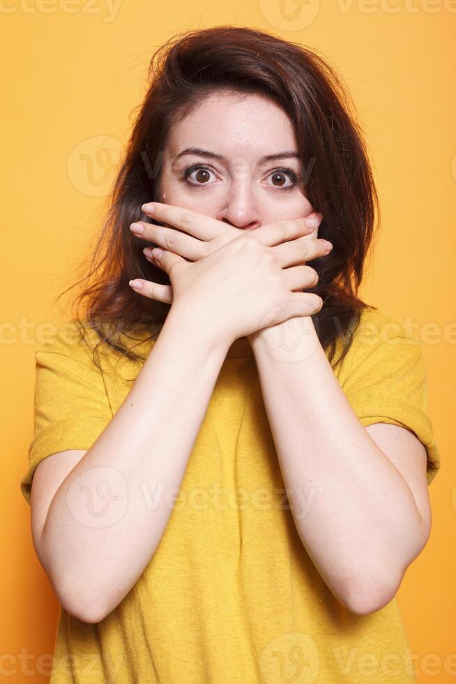 Shocked woman with her mouth covered in both hands as she stares at an unbelievable situation. Concerned female person stands against orange background, looking both surprised and terrified. photo