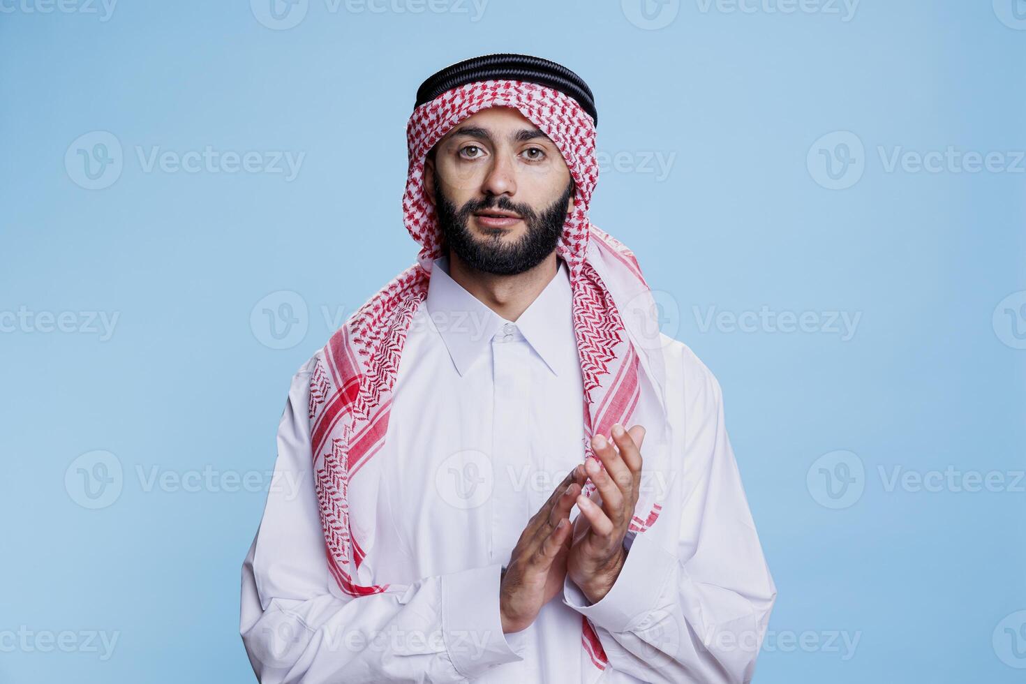 Muslim man in thobe and ghutra applauding with hands, showing support and approval studio portrait. Arab person wearing traditional outfit clapping arms and looking at camera photo