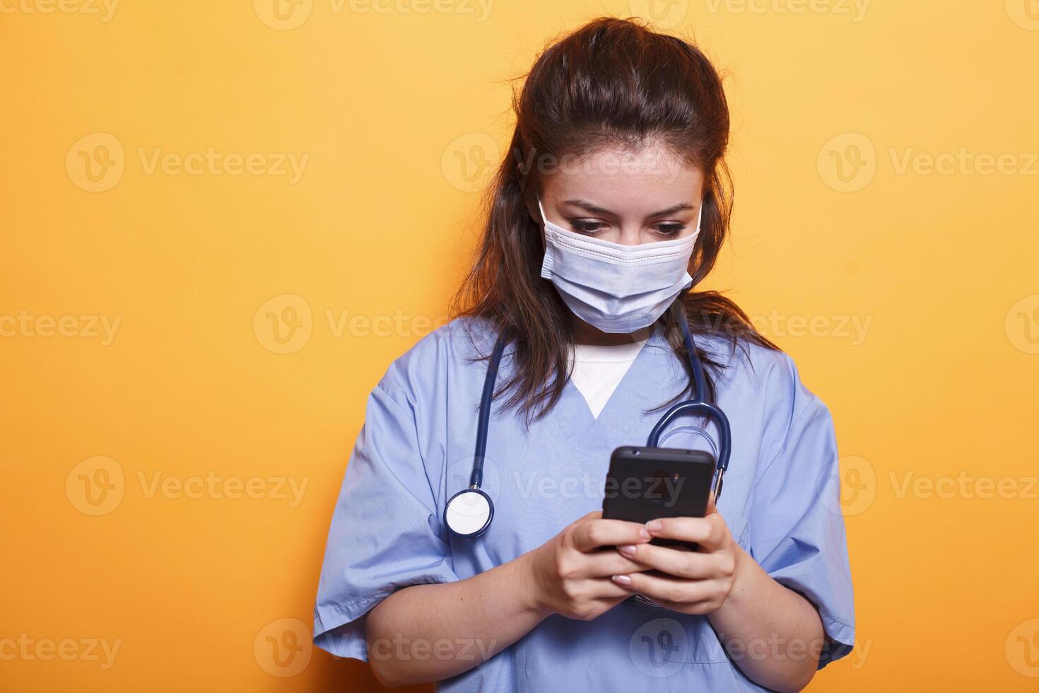 Caucasian female doctor utilizing modern technology while wearing face mask against coronavirus. Medical assistant with stethoscope texting on her mobile phone during pandemic. photo