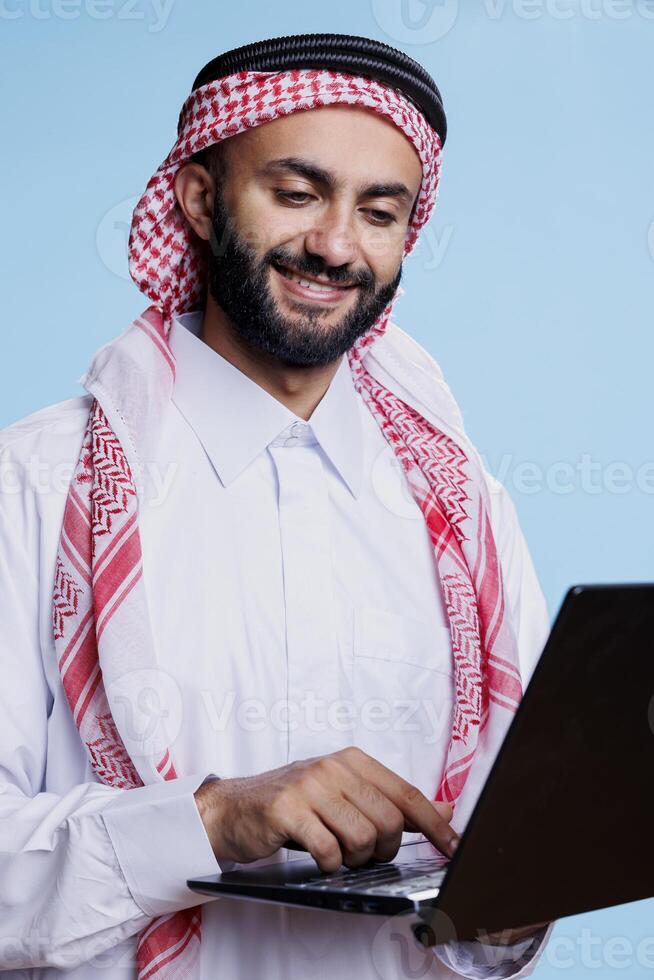 Smiling muslim man holding laptop and pressing button on keyboard with cheerful expression. Arab person wearing traditional clothes typing and sending message on computer photo