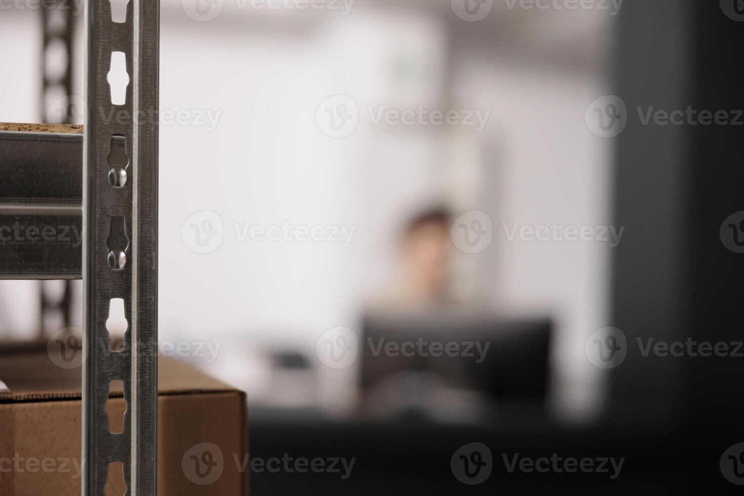 Selective focus of warehouse shelves full with cardboard boxes, in background employee working at counter desk, checking clients online orders. Storehouse supervisor preparing packages for delivery photo