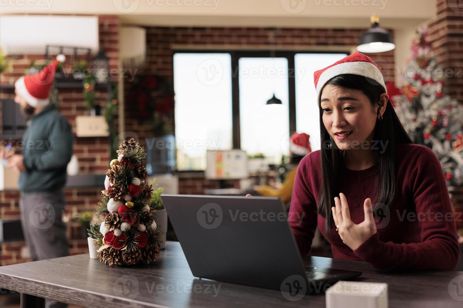 Asian woman in santa hat chatting in online meeting with remote coworker in festive office during christmas eve celebration. Employee discussing project in videocall at decorated workplace photo
