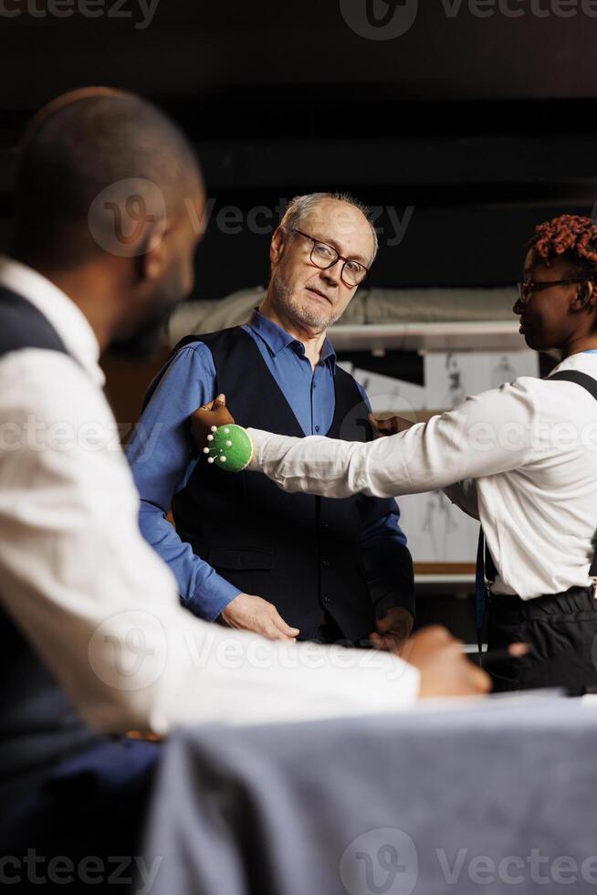 Client trying on premium quality suit vest under the expert detail oriented suitmaker eyes. Happy assistant seamstress proud by finished bespoke sartorial piece comissioned by senior customer photo